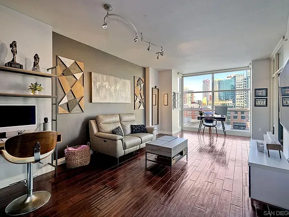 Living room in California with dark wood floors, gray accent wall, tan sofa, large windows, abstract art, and shelves.