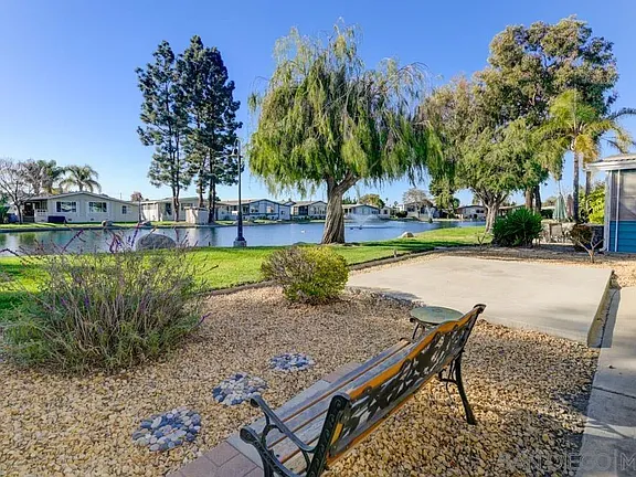 Park with rustic bench on pebbled ground, pond, trees and houses.