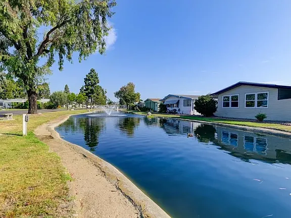 Blue water, single-story house, trees, fountain, and bright sky.