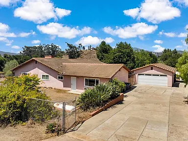 Single-story pink California house with tiled roof, separate garage, paved driveway, and surrounding shrubs and trees.