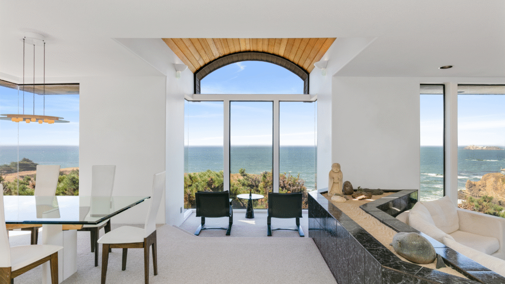 California modern living room with large windows, ocean view, glass dining table, chairs, and a stone sculpture.