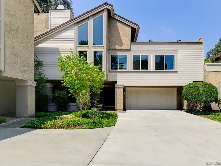 neighborhood house with angled roof, large windows, beige siding, driveway, and green trees.