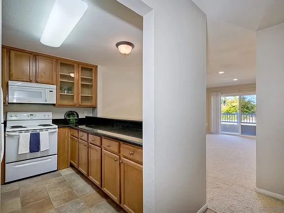 Kitchen in California with wooden cabinets, black countertops, white stove, microwave, and adjacent living room.